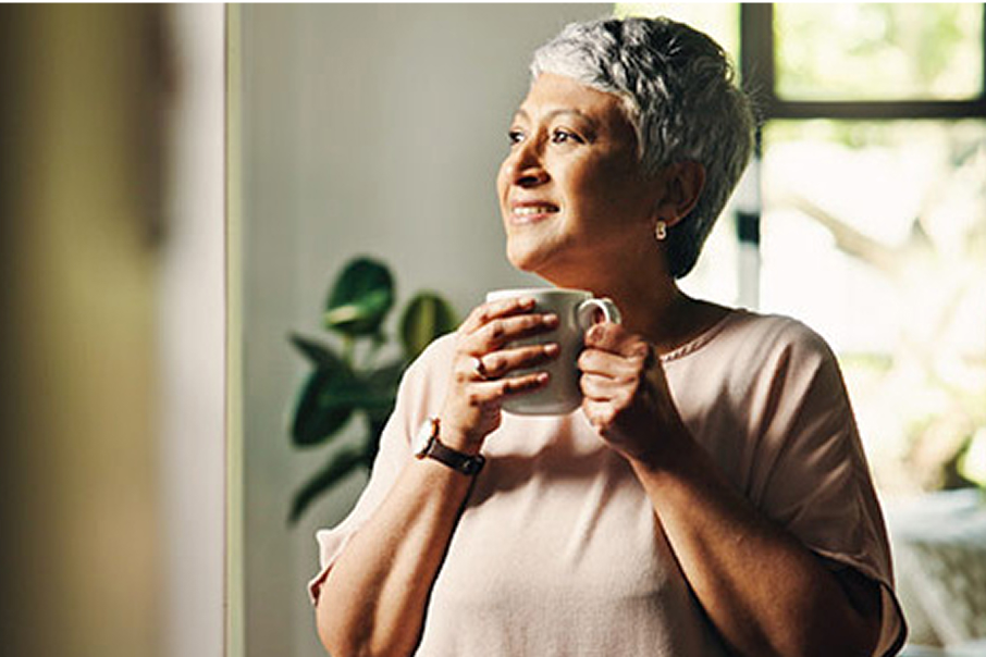 A smiling middle-aged woman looks out a living room window with a coffee mug in hand.