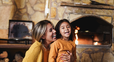 Child is being embraced by a parent while they sit on the floor in front of a lit fire in the fireplace.
