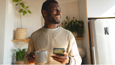 A smiling man holds a coffee cup in one hand and his phone in the other as he looks off in the distance.